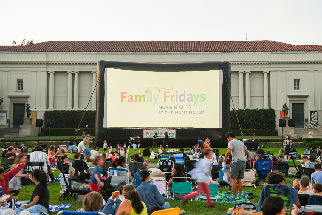 A crowd of people sits on a lawn in front of a large inflatable screen reading "Family Friday: Movie Nights at The Huntington." A large white building with columns and a red roof is in the background.