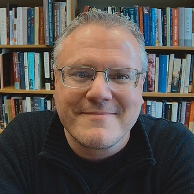 A smiling person wearing glasses, in front of a book shelf.