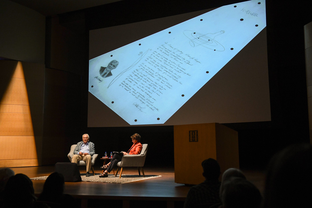 Two people sit on a stage in front of an audience, with a screen displaying a poem.