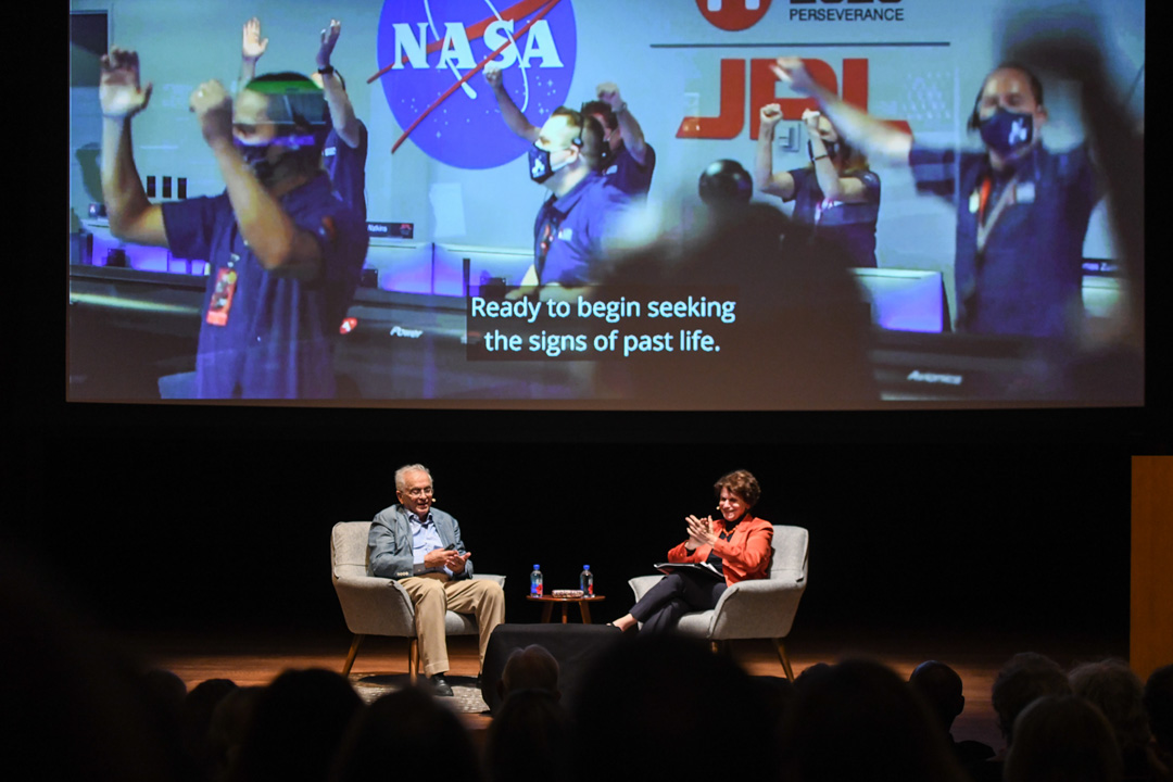 Two people sit on a stage in front of an audience, with a screen displaying a video of a NASA control room.