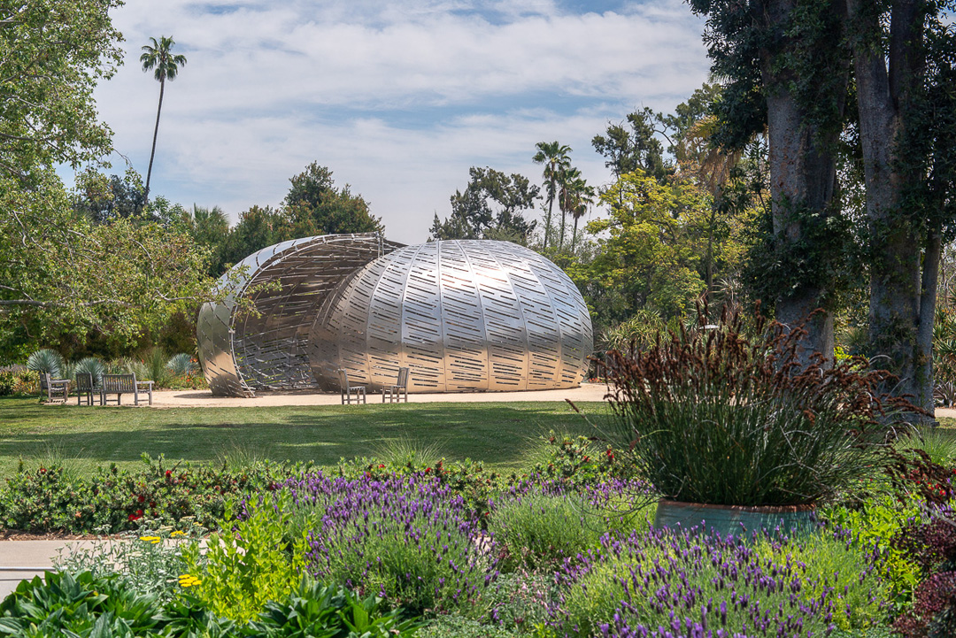 An aluminum sculpture, surrounded by a lawn and a lush garden.