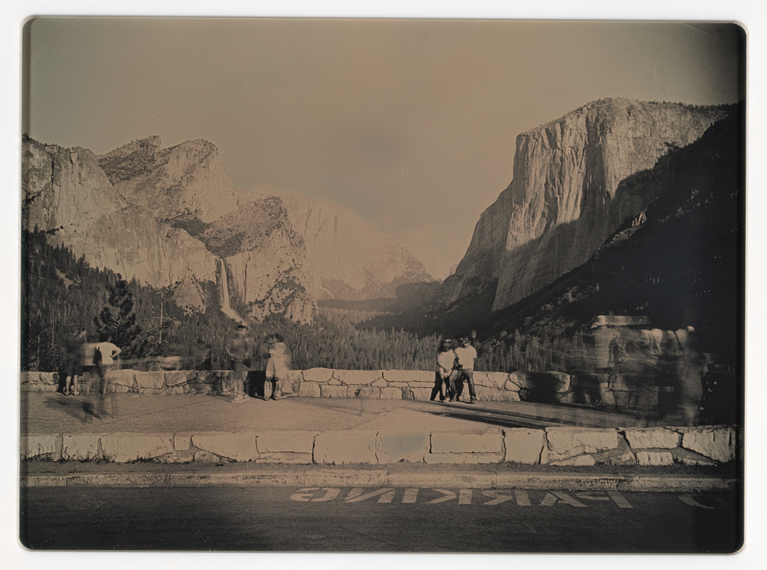 A grayscale image of people near a paved road in a national park with large mountains and a forested valley..