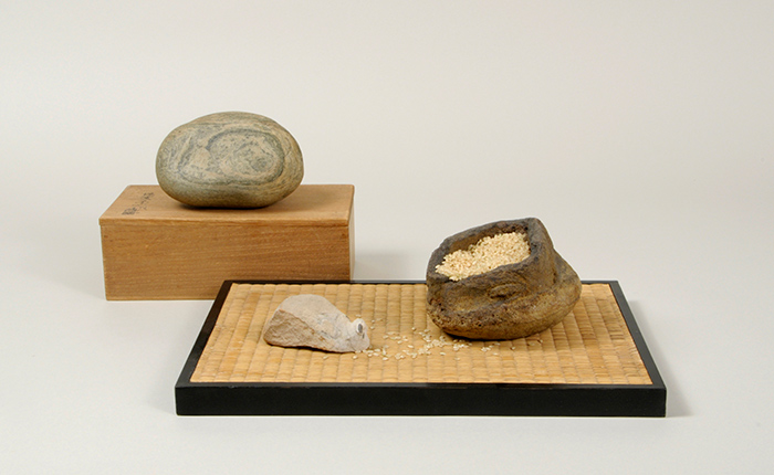 Two wood pedestals display three stones, one with an impression of a sleeping cat, one that resembles the shape of a mouse, and one that appears to be a bag filled with grain.