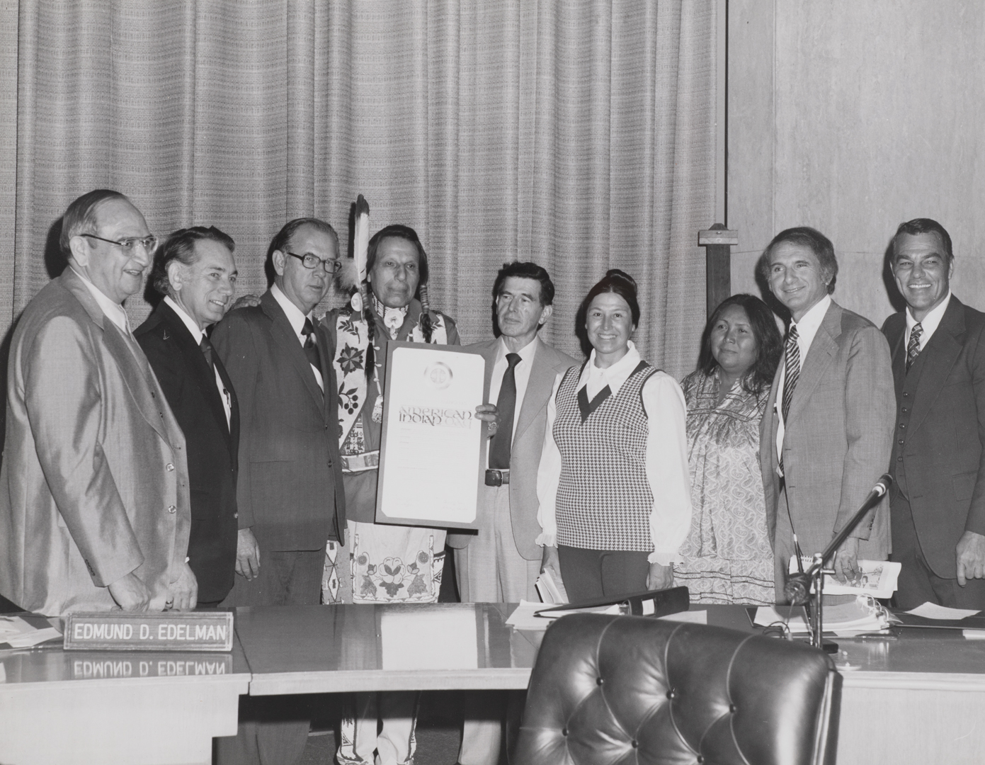 A black-and-white photo of a group of people behind a desk; one person holds a document.