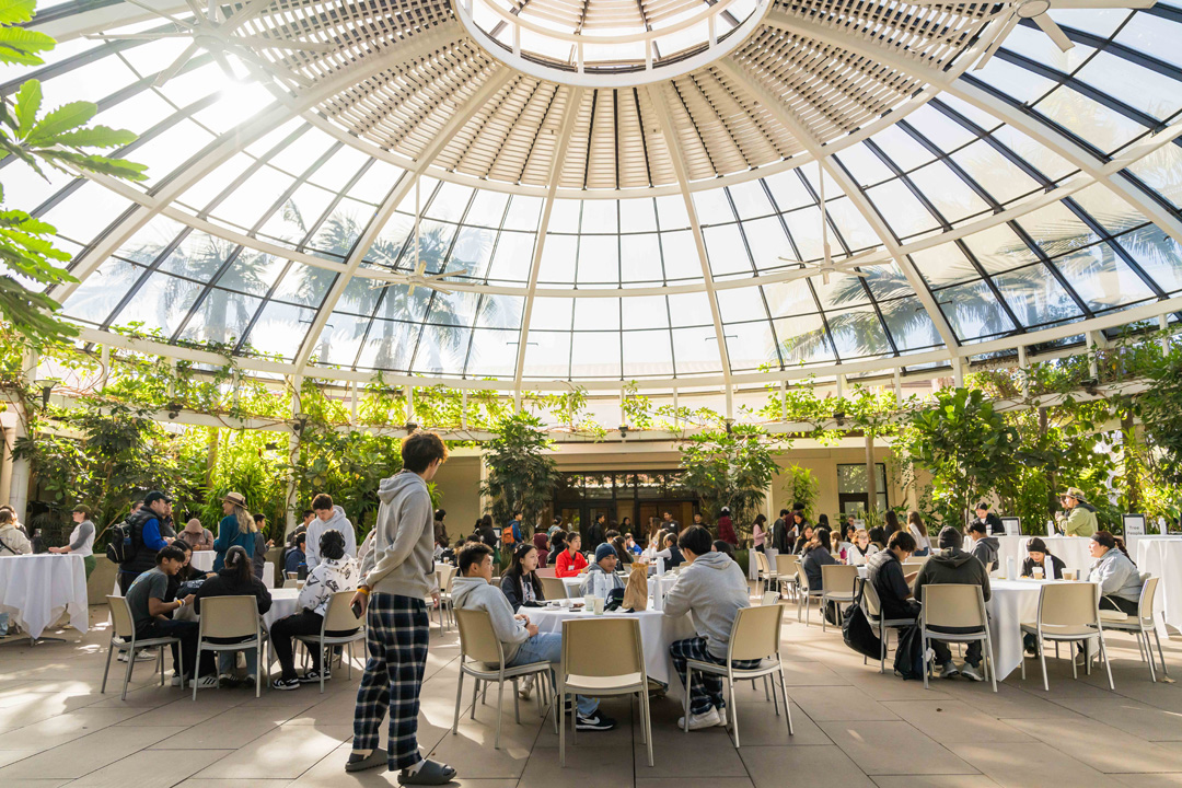 People sit at tables under a glass dome filled with plants.