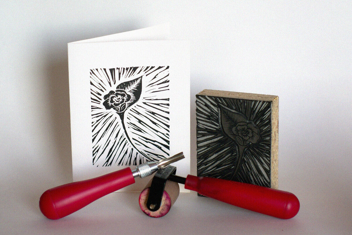 A linoleum block, carving and printing tools, and a folded paper with a black ink print of a flower.