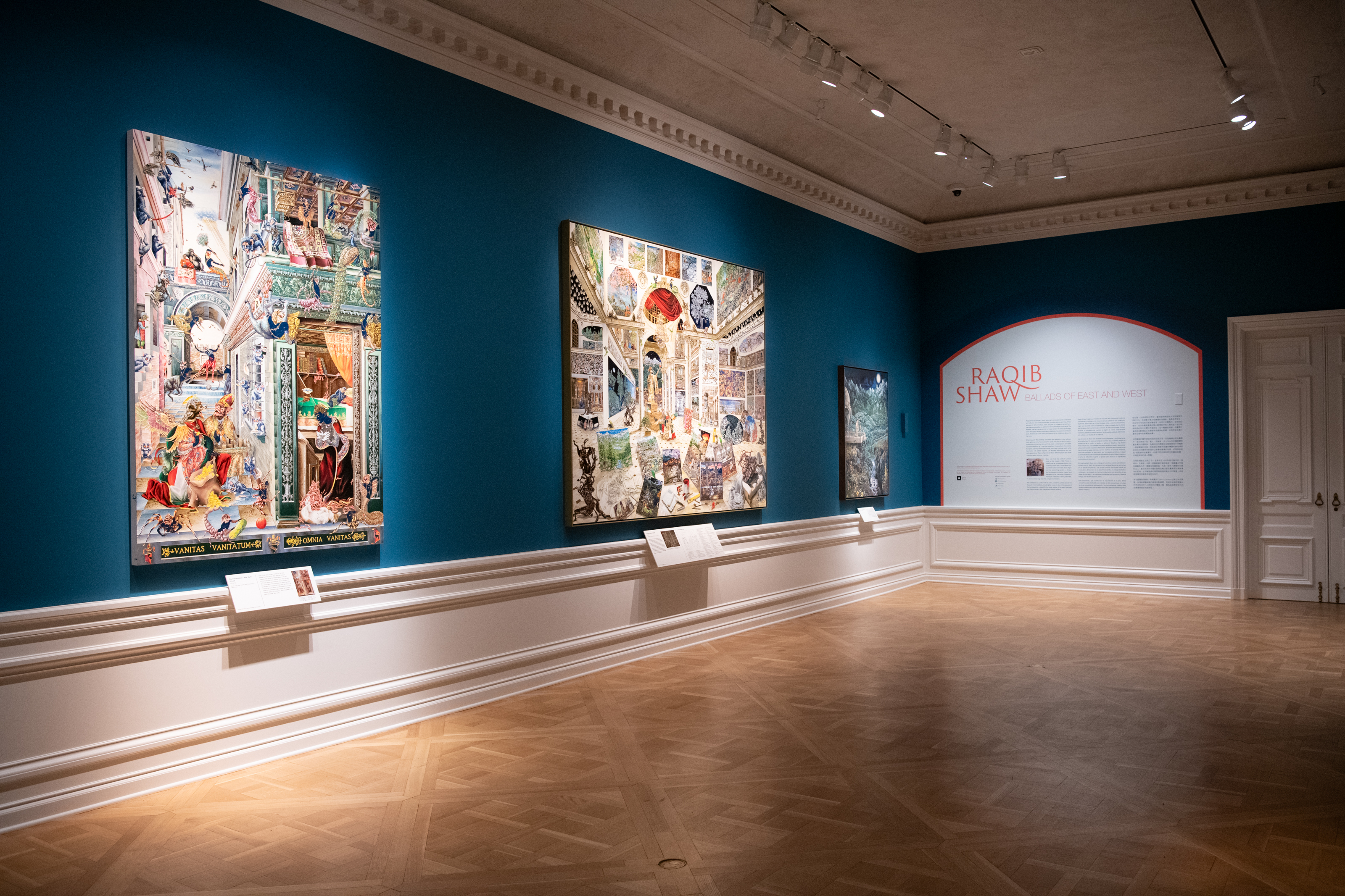 Two walls in a gallery with blue walls, white trim, and wood floors; three large artworks hang in a row on the left-side wall, and a block of introductory text is displayed on the right-side wall.