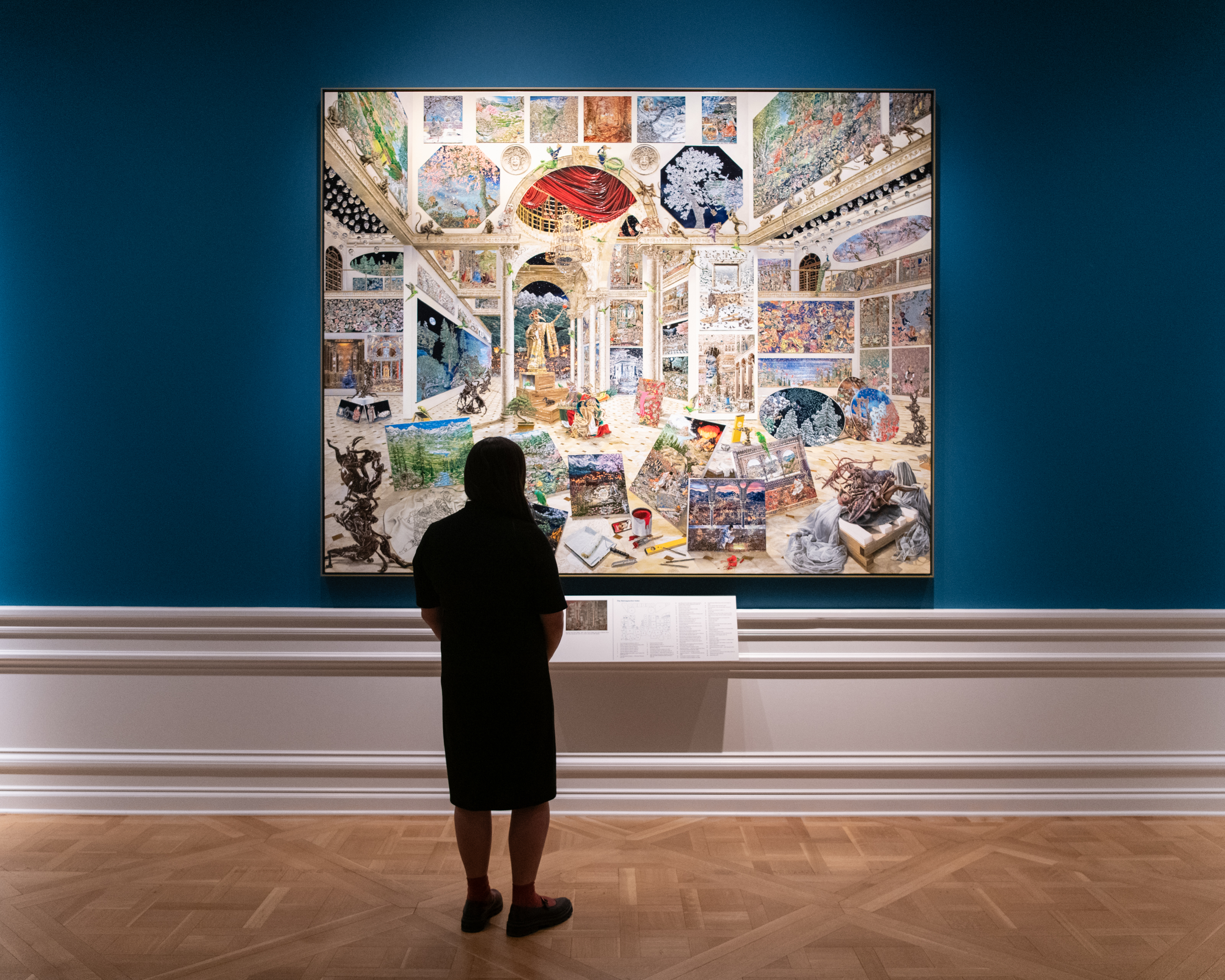 A person in shadow stands in front of a large, vibrant painting depicting an opulent and cavernous room filled with artworks.