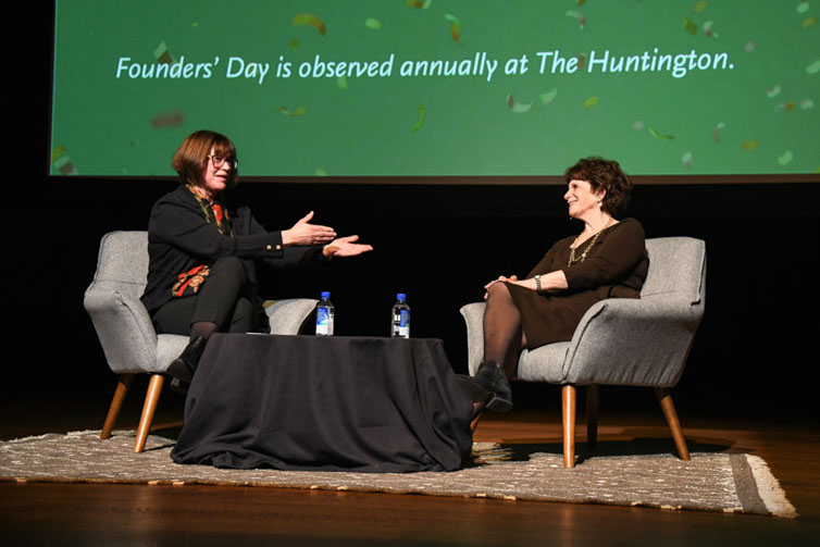 Two people sit on chairs on a stage in front of a green screen.