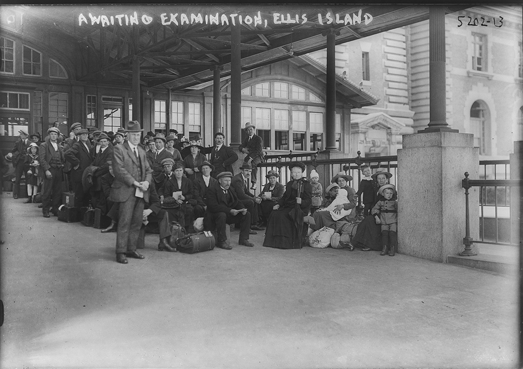 A black and white photograph of a large group of people under the awning of a building with large windows. White writing, near the top of the print, reads "AWAITING EXAMINATION, ELLIS ISLAND, 5202-13."