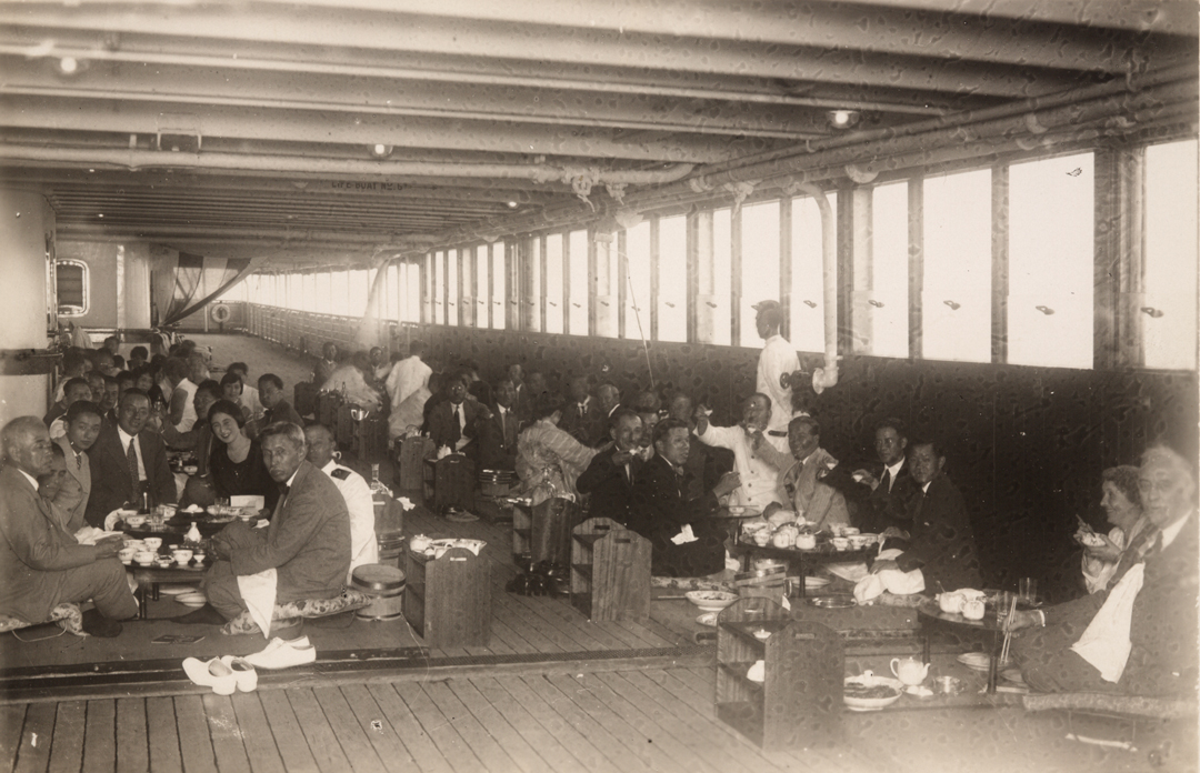 A black-and-white photo of people seated on the floor around a low table on a boat deck.