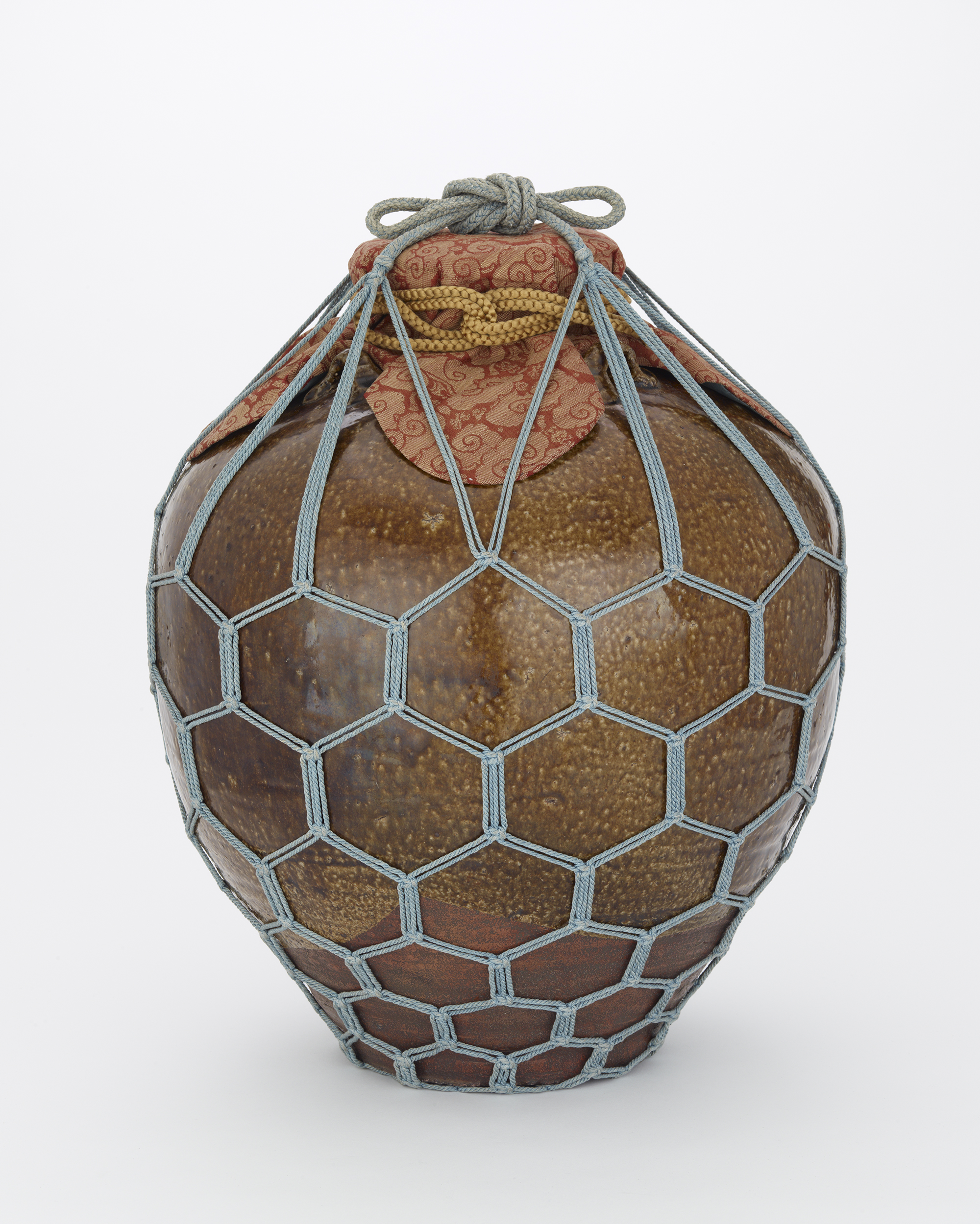 A brown ceramic jar with a patterned cloth over the lid; the object is wrapped in a gray netting with a hexagonal pattern.