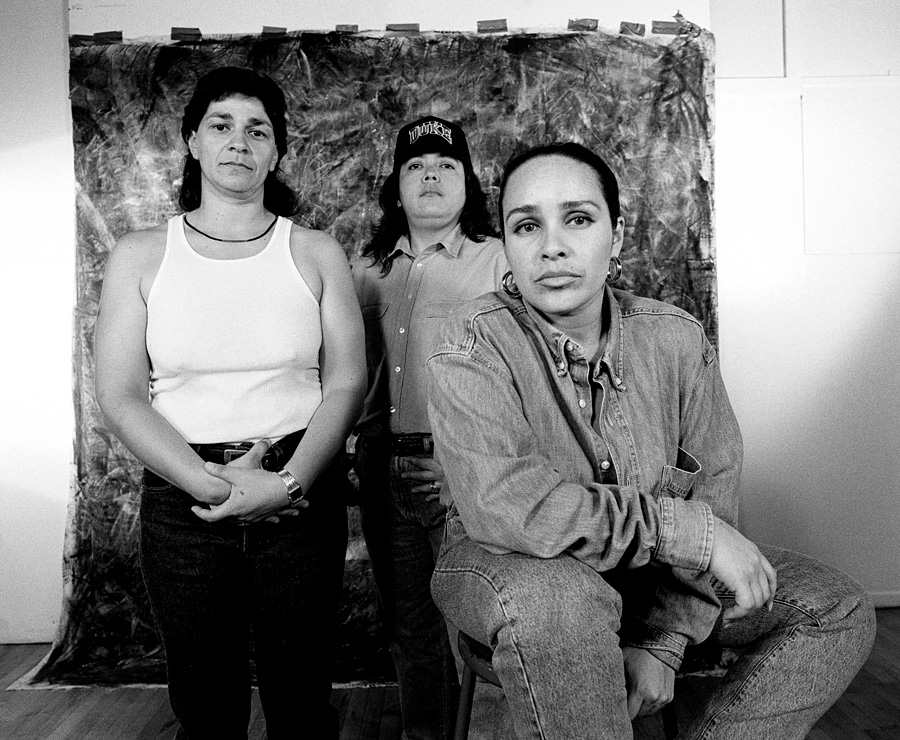 Group of three queer women posing for a portrait in front of a roughly textured backdrop