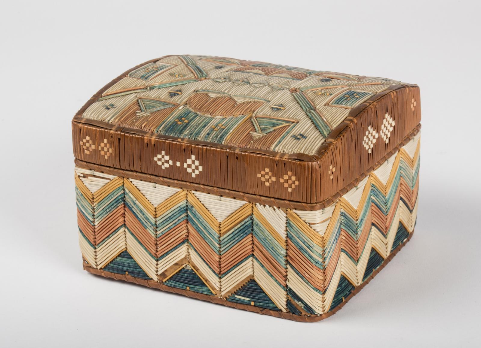 Angled view of a small colorful box with a lid, decorated with lengths of porcupine quills forming a geometric design on the lid and chevron patterns on sides.