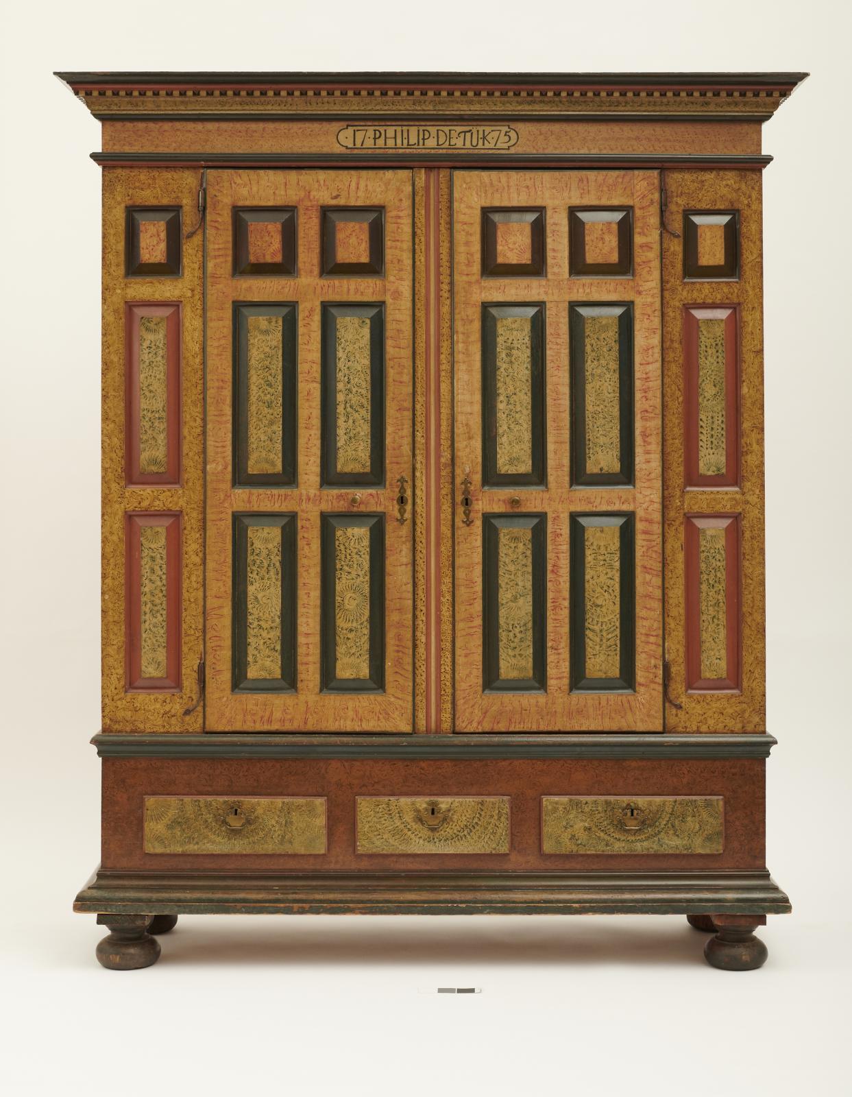 Large wooden wardrobe with panels, each with a border of red, green or black and painted decoration simulating the look of wood grain; drawers across bottom and cornice at top.