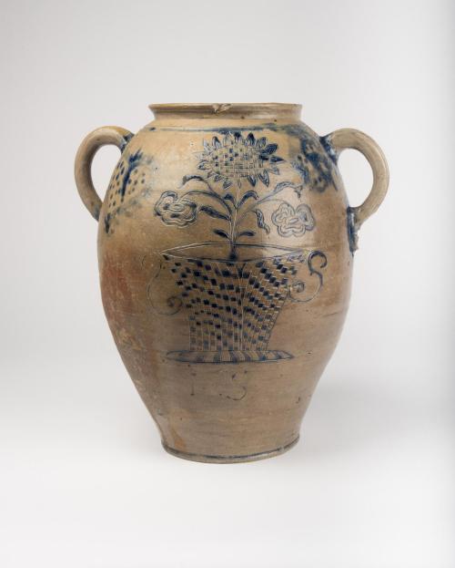 Front view of ovoid jar with handles near the top, decorated with blue pigment and incised decorations, with a flower in a checkered pot at the center and the initials “IS” below.  