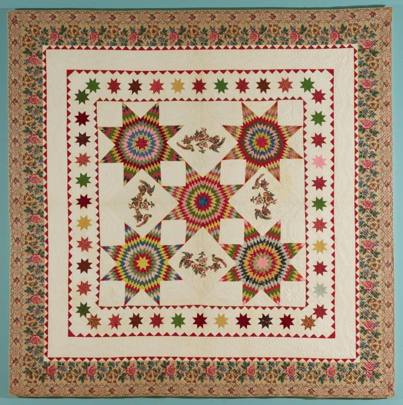 Elaborate fabric quilt featuring an arrangement of five star shapes in the center, surrounded by a border of smaller star shapes and an outermost border of appliqued fabric in a floral pattern.