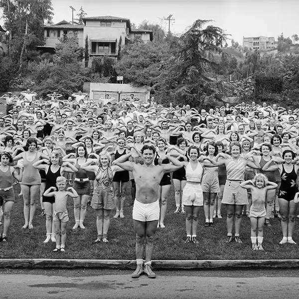 A group of 100 or so people in bathing suits and shorts, make a "crab" pose, showing off the muscles in their arms. 