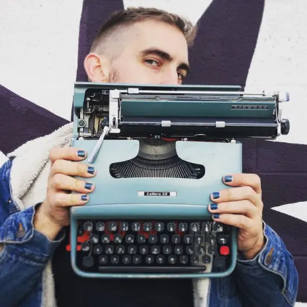 A person with blue fingernails holds up a typewriter in front of their face.
