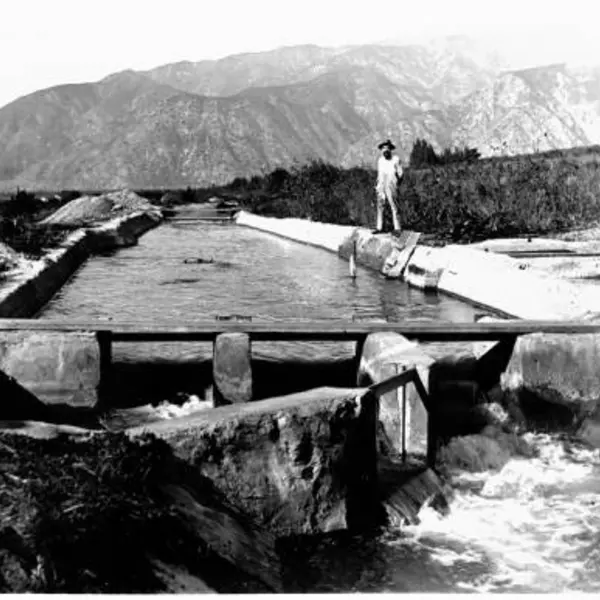 Black and white photo of man standing on side of irrigation canal full of flowing water.