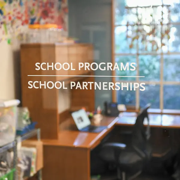 An open laptop is set on a desk seen through a glass window that says "School Programs" and "School Partnerships."