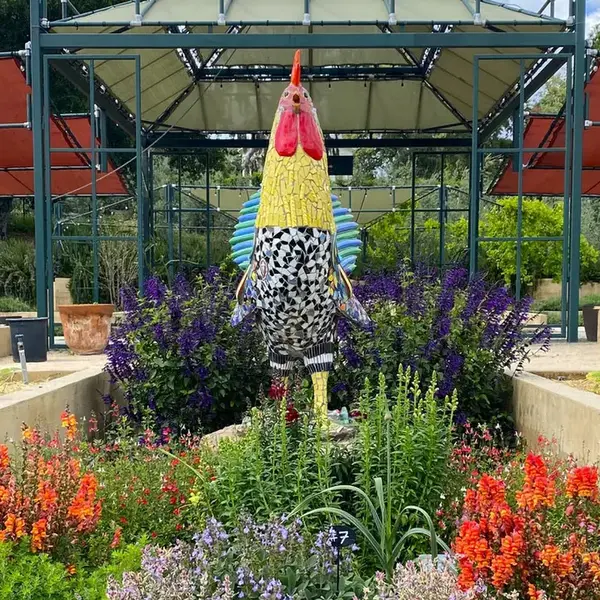 A large rooster sculpture made of colorful tiles in a garden of flowering plants.