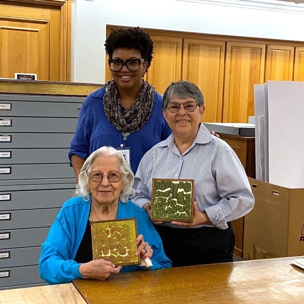 Three people stand together smiling, holding two tiles.