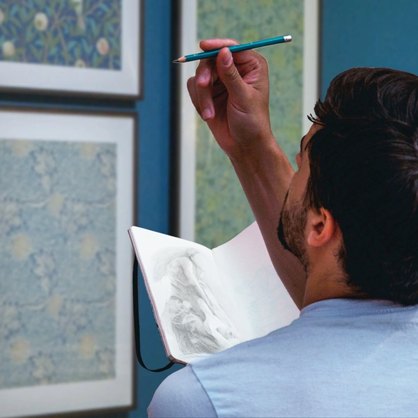 A person holds a sketchbook and pencil while looking at artwork in a gallery.