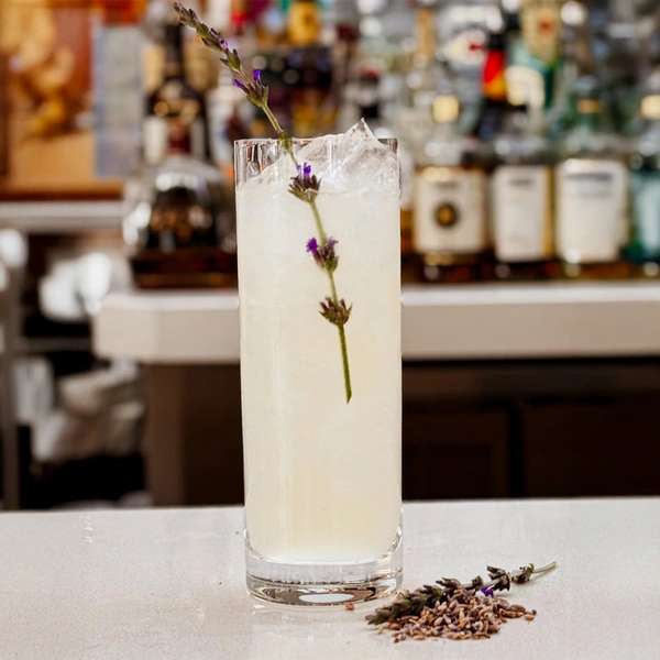 A tall narrow glass filled with light-yellow lemonade and a sprig of lavender.