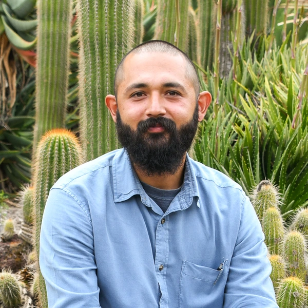 A person with short hair, a full beard, wears a blue shirt in a green garden.