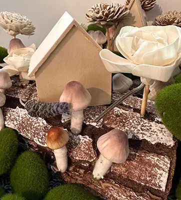 A miniature "fairy house" display with an "A" frame home and a fabric rose as a tree.