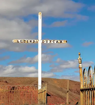 A large white metal cross with the name "Lucinda Duncan" in black lettering on the horizontal beam. A cloudy blue sky and orange desert landscape can be seen in the background.