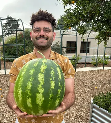 A person stands in a garden holding a watermelon.