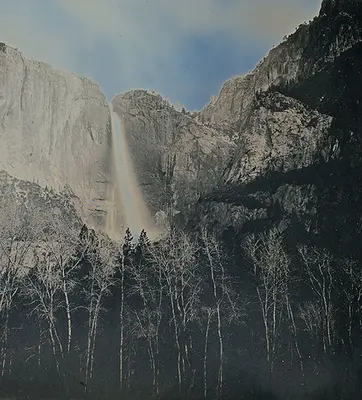 A waterfall cuts through a crevice in a tall mountain range, disappearing behind a forest of trees.