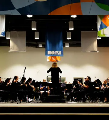 A conductor stands in front of an orchestra on a stage with colorful banners.