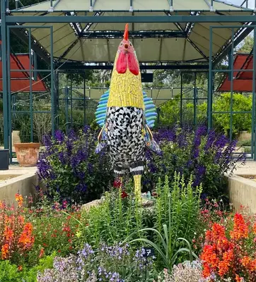 A large rooster sculpture made of colorful tiles in a garden of flowering plants.