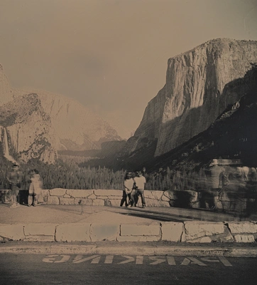 A grayscale image of people near a paved road in a national park with large mountains and a forested valley.