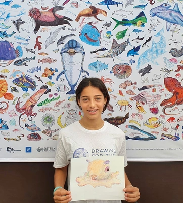 A young person holds a drawing on paper while standing in front of a poster filled with colorful illustrations of sea creatures.