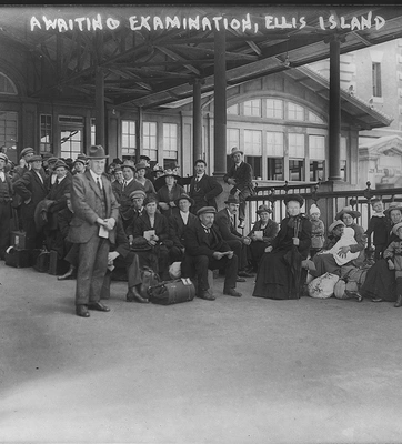 A black and white photograph of a large group of people under the awning of a building with large windows. White writing, near the top of the print, reads "AWAITING EXAMINATION, ELLIS ISLAND, 5202-13."