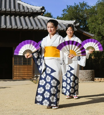 Two people in colorful Japanese outfits hold fans in front of a building in a garden.