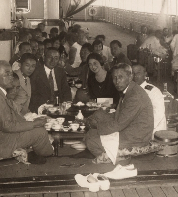 A black and white photo of people seated on the floor around a low table on a boat deck.