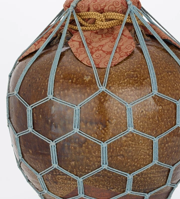 A brown ceramic jar with patterned cloth over the lid, the entire object is contained within a secure gray netting with a hexagonal pattern.