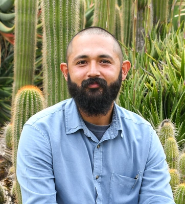 A person with short hair, a full beard, wears a blue shirt in a green garden.