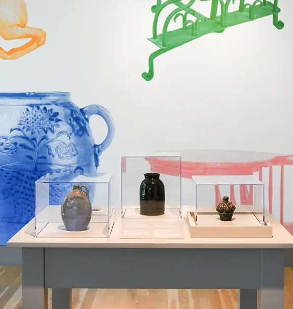 Stoneware jugs sit on a small table in front of a wall painted with a blue jug, red chair, and other colorful items.