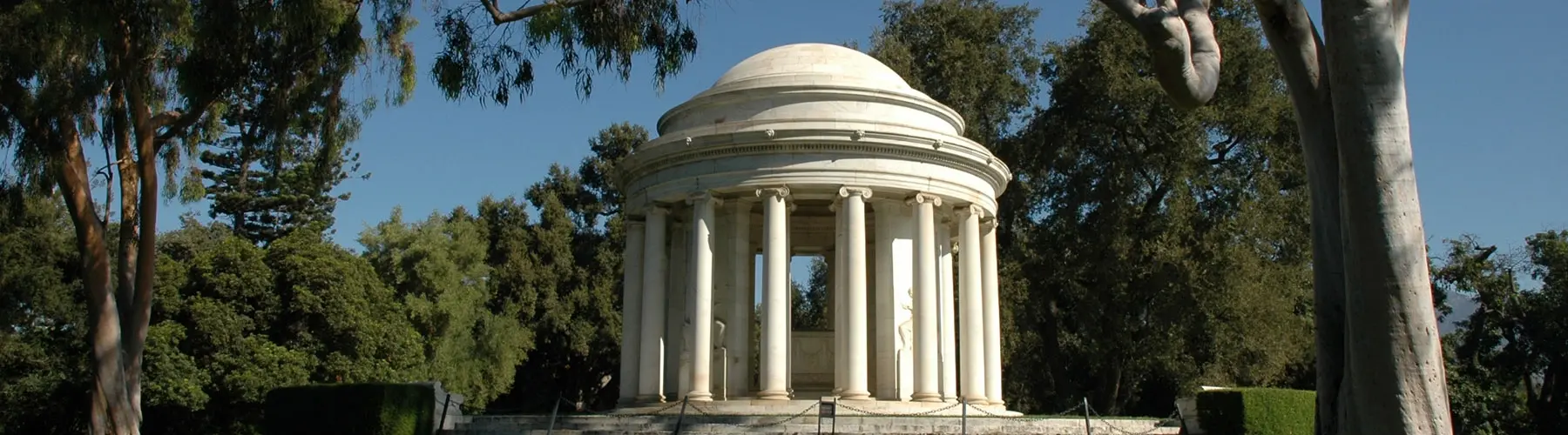 A marble rotunda designed in the form of a Greek temple envelopes an ornate sarcophagus. A series of hanging black chains some 2-3 feet off of the ground lines the foreground of the rotunda.