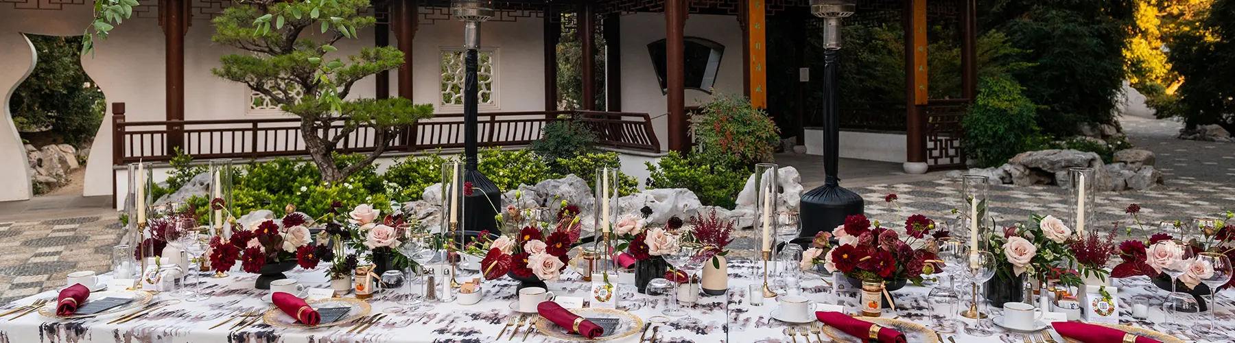 A dining table with place settings of red and light pink set up in an outdoor courtyard.