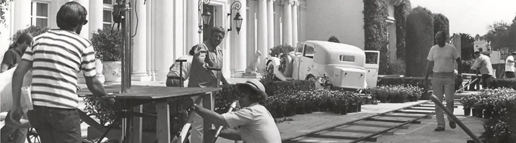 Black and white photo of a car crashed in front of a mansion with filmmakers and camera dolly tracks in front.