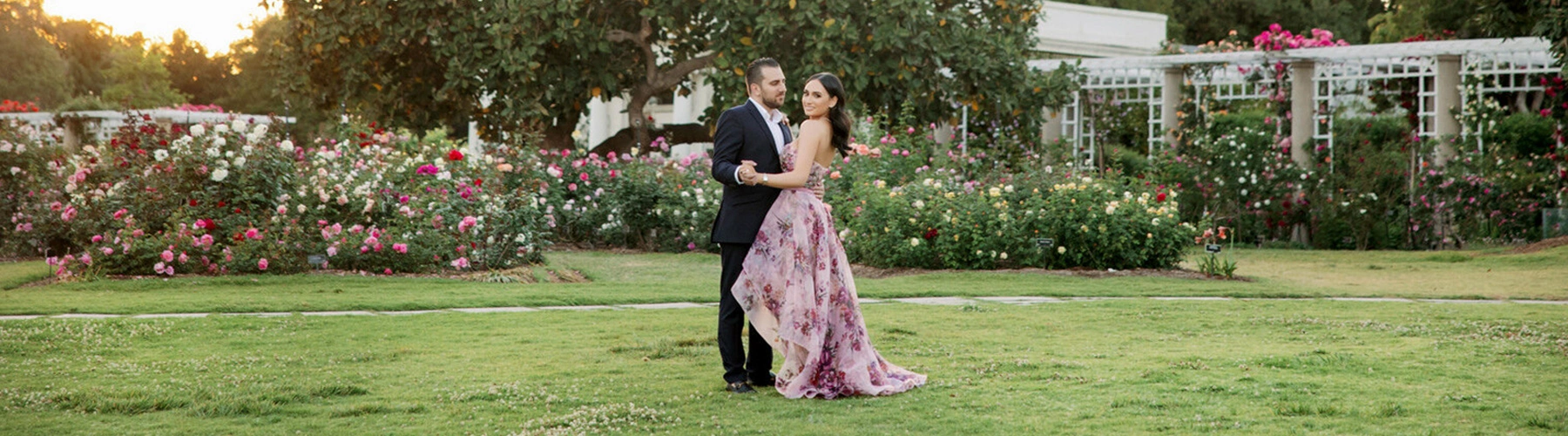 An engaged couple pose in front of beds of blooming roses.