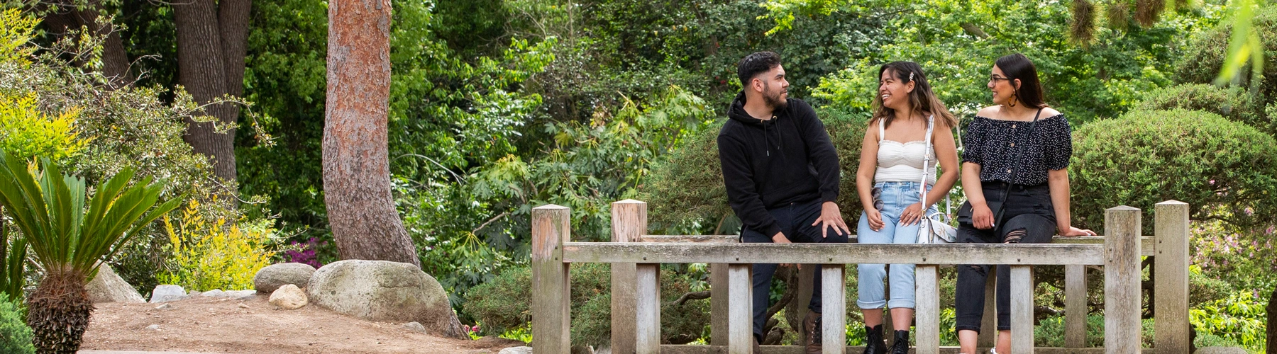 Three young people sit on a low bridge over a creek in a verdant Asian-style garden.