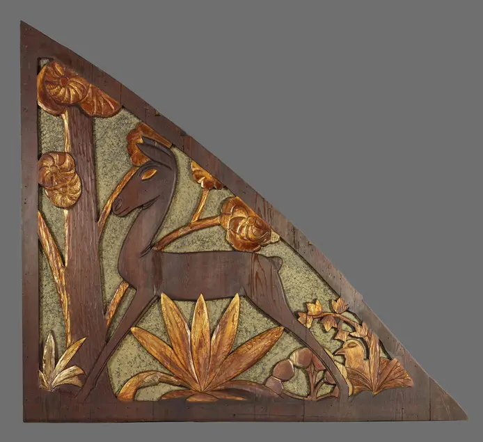 Carved wood triangular panel. The panel includes a deer with gold eyes and a gold bird on the ground. The background of the panel has gold trees and grass and flowers made out of wood. 